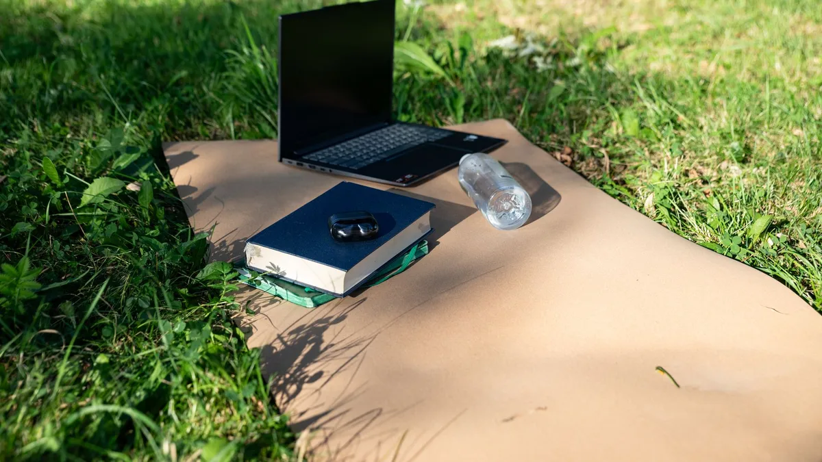 Laptop, books, and water bottle set on a mat in a sunny outdoor setting, ideal for remote work.